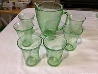 Full set of six green uranium glass water glasses arranged in a semicircle with the matching pitcher behind, all on a white surface showing the translucent green glass and the delicate etched floral pattern.