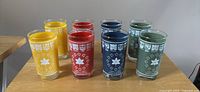 Photo showing set of 8 tall commemorative glasses in yellow, red, blue, and green. Each glass features white provincial crests along rim and central maple leaf designs.