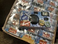 Disposable film cameras arranged in original cardboard box with one camera placed on top showing front design with camera lens, flash, and beer branded bottle cap graphics.