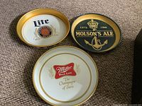 Three metal beer trays arranged together showing fronts: Molson's Ale, Miller High Life, and Lite designs with gold rims.
