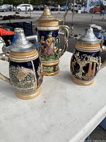 Photo showing all three beer steins, two shorter navy with beige decorations and one taller colorful musical stein with pewter lids