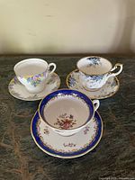 Three teacups and saucers displayed together on a marble surface showing their unique floral patterns and gold trims.
