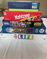 Stack of 5 boxes: Painted Hills 500 piece puzzle on top, Yahtzee box second, Motorecords 1000 piece puzzle third, White Mountain 500 piece puzzle fourth, The Game of Life board game bottom.