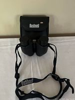 Front view of black Bushnell binoculars with neck strap and carrying case, placed on white surface