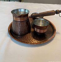 Copper Turkish coffee set on white fabric background, showing cezve with wooden handle, two cups, and round hammered copper tray from side angle.