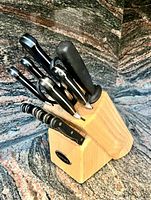 Front and side view of 13 knives and scissors with black handles in a light wooden knife block.