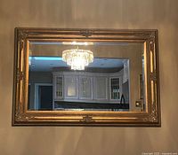 Front view of large rectangular mirror with ornate gold wooden frame reflecting a chandelier and part of a kitchen.