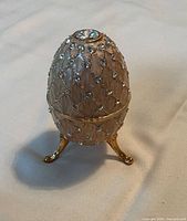 Front view showing decorative egg standing on three metal legs, enameled surface with clear rhinestones.