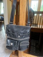 Front view of black leather crossbody bag showing all front pockets including one with zipper, one with button closure, and two smaller zipper pockets, hanging from an adjustable strap.