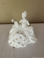 Front view of the white porcelain figurine of a seated woman with gold accents on dress, arm bands, and shoes.