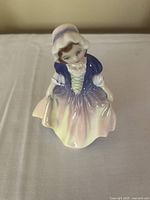 Front view of Royal Doulton small bone china figurine of a girl wearing a bonnet and multicolored dress.