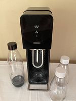 SodaStream carbonating machine with three bottles: one 1L bottle with black cap and two 500ml bottles with white caps, shown on a white surface.
