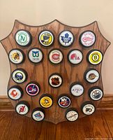 Front view of wooden shield plaque adorned with 22 vintage NHL hockey pucks featuring team logos such as North Stars, Blues, Maple Leafs, Red Wings, Blackhawks, Whalers, Canadiens, Bruins, Flames, Jets, Penguins, Islanders, Kings, Oilers, Capitals, Flyers, Rangers, and NHL logo puck centered.