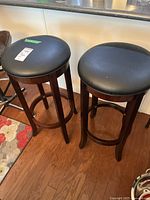 Two matching wooden bar stools with black round cushioned seats placed side by side on wooden floor near kitchen counter.