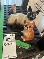 Photo showing both Beswick cat figurines on a black plastic surface with auction tags; visible crack on the Siamese-style cat figurine.