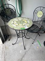 Bistro set showing round mosaic tile top table and two different folding metal chairs with decorative backs.