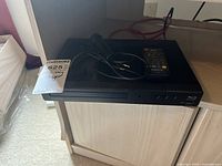 Front view of the Sanyo FWBP507FF Blu-ray player with a remote and microphone placed on top of it, showing disc slot and buttons.