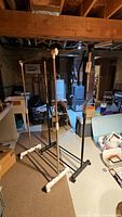 Photo showing two standing clothing racks made of black metal rods with white plastic connectors and feet, in a basement setting with other household items in the background.