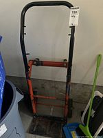 Front view of orange and black metal dolly with wheels and foldable base plate against garage wall.