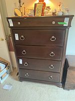 Front view of a tall wooden dresser with five drawers and metal round ring handles. Shows dark wood finish and solid base.