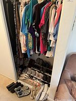 Closet with hanging ladies shirts and pants in various colors and patterns, shoes placed on shoe racks and floor, shoe stretchers and shoe stands visible