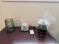 Three tabletop lamps on a wooden surface: green ribbed glass lamp, clear plastic bead lamp, white fibre optic lamp.