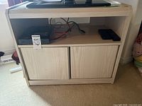 Full view of the media unit with light wood finish, showing open shelf with cables and remotes, and closed cabinet doors below.