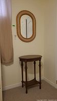 Photo of oval wicker style mirror mounted on wall above vintage wooden side table with curved legs and lower shelf.