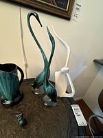 Photo showing two green bird figurines with long curved necks, a tall white bird figurine with a curved neck, small ceramic animal figurine, and a green glossy pitcher. Items are placed on a table with intricate carvings.