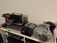 Full view of the lot on a shelf showing multiple electronics including boombox, radios, VCR, VHS tapes and rewinder.