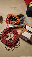 Plastic container holding a mix of black power cords and orange extension cord coiled, with a thick red extension cord coiled on the floor nearby.