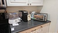 Image showing five small appliances on kitchen counter: food steamer, Keurig coffee maker, can opener, electric kettle, toaster oven.