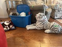 View of large white tiger stuffed animal lying on wooden floor next to sofa and blue storage bin containing various smaller stuffed animals, a rubber chicken, and cushions.