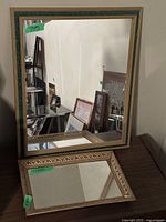 Rectangular gold framed wall mirror standing behind rectangular mirrored bottom dresser tray on wooden surface.
