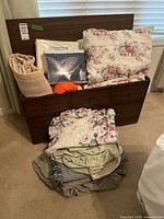 Full view of the wood trunk with blankets and linens stacked on and beside it, includes a plastic wrapped framed item and orange yarn.