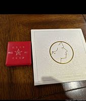 Red USSR coin case with inscriptions 1945-1975 and CCCP alongside white UK coin booklet with gold embossed profile of Queen and text.