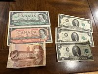 Photo showing three US $2 bills from 1976, 1967 Canadian $1 bill, 1954 Canadian $2 bill, and 1986 Canadian $2 bill on a wooden surface.