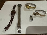 Photo showing five vintage ladies watches on a white surface, varied styles and bands including stretch metal bands and decorative elements.