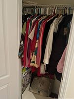Wide view of closet displaying multiple women's shirts, jackets and sweaters on hangers with additional folded sweaters inside a tote bag on floor.