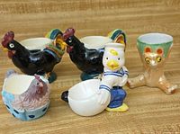 Group view of five ceramic egg holders: two roosters, hen basket, sailor duck, cat cup