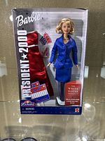 Front view of the boxed Barbie doll with blue suit and accessories, showing included red dress and campaign buttons.