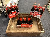 20 vintage Coca-Cola glass bottles arranged in carriers and loose in a wooden tray, displayed on carpeted floor.