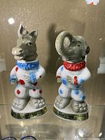 Front view of both ceramic whiskey bottles shaped as bear and elephant in clown costumes.