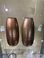 Front view of two hammered copper color metal vases side by side on glass shelf, reflecting light.