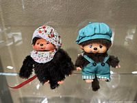Both Monchhichi dolls on clear surface, showing full front and side profile with clothing details