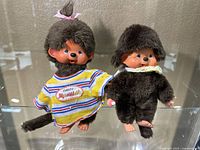 Photo showing 2 Monchichi plush dolls, one in yellow striped shirt, the other in brown fur outfit.