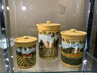 Three lidded ceramic containers displayed, showing the painted rural landscape and yellow accent on rims and lids.