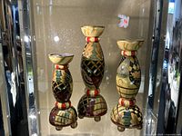 Three wooden candle sticks with detailed hand-painted floral and geometric designs displayed upright in a glass cabinet.