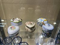 Front view of 5 small lidded containers showing heart-shaped and round ceramic and porcelain pieces with floral and bird decorations.