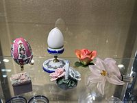 Overview of all six small decorative items displayed on glass shelf including two egg-shaped ornaments and four floral figurines.